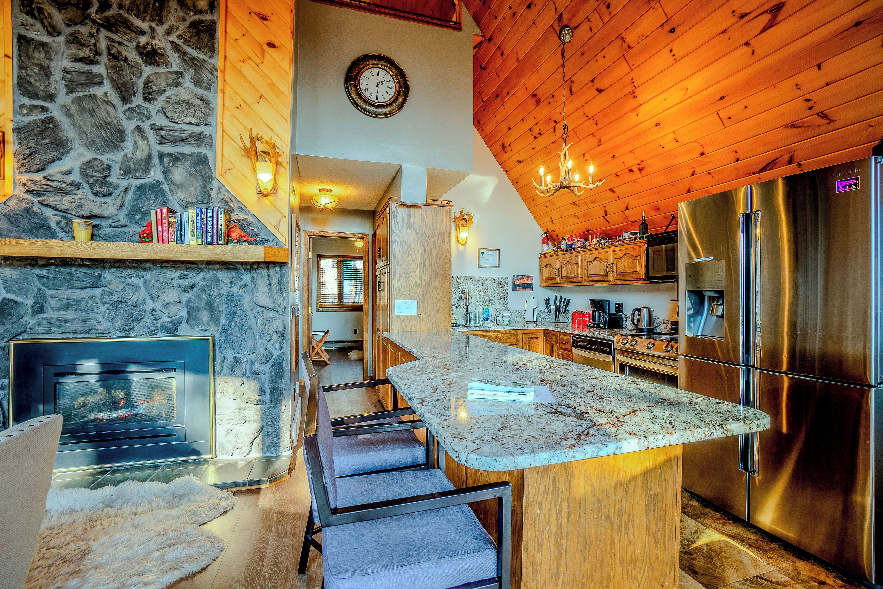 A modern kitchen with a stone fireplace, wooden ceiling, and stainless steel appliances.