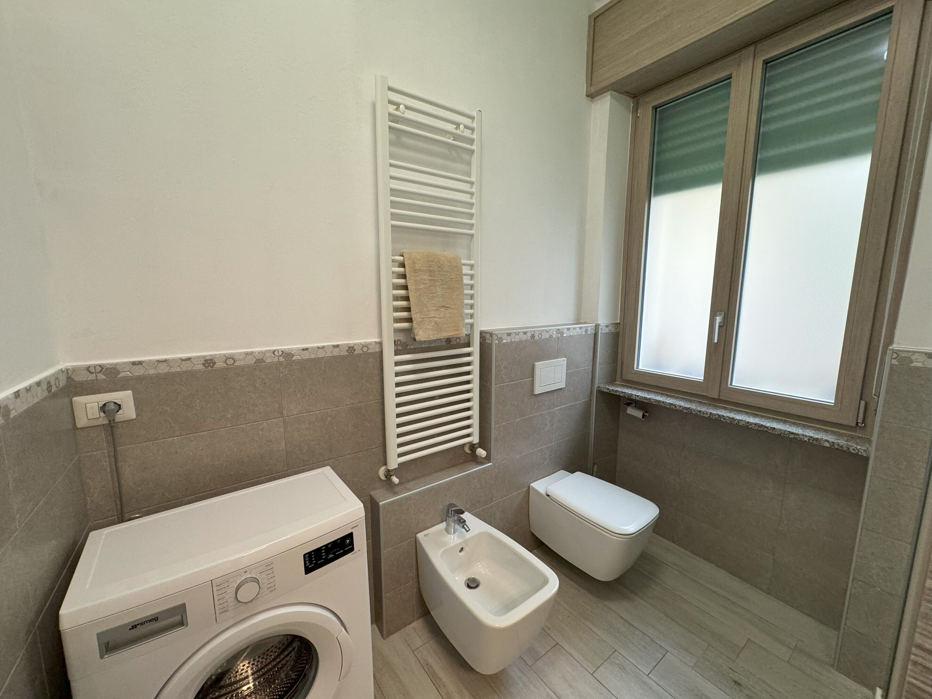 A bathroom with a washing machine, bidet, and a window with frosted glass.