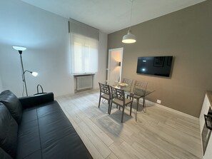 A modern living room with a black leather sofa, a glass dining table with chairs, a floor lamp, a radiator, and a TV mounted on the wall. - Am'Arena Apartment - Sleeps 4, 300 meters from the Arena (Verona)