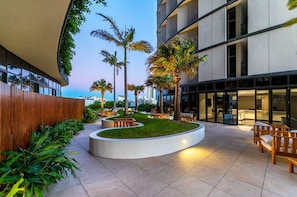 A modern outdoor area with a curved seating arrangement, palm trees, and a building with large windows. - Rooftop Infinity Pool! 5 Guests - Free Park & Gym (Fortitude Valley)