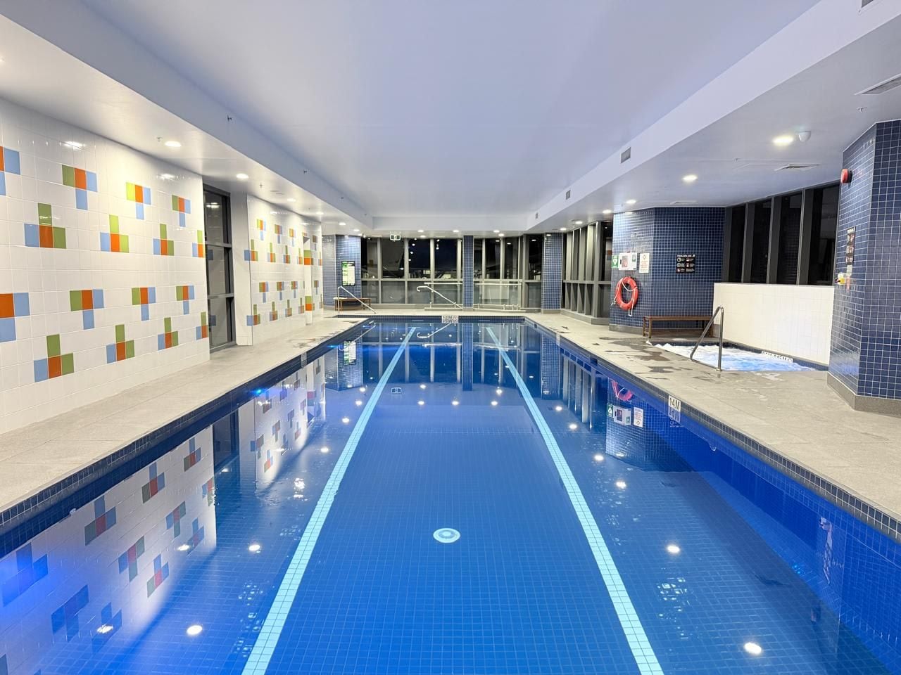 Une piscine intérieure avec des couloirs bien délimités, un poste de surveillance pour le sauveteur et un mur carrelé orné d’un motif géométrique.
