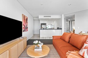 A modern living room with a flat-screen TV, a round coffee table with a book and flowers, a sofa, and a kitchen in the background. - River Views from Level 43 | Free Parking, Pool & Gym (Brisbane City)