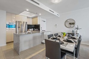 A modern kitchen with stainless steel appliances, a dining area with a white table set for four, and a round mirror on the wall. - City Skyline from Level 37 | Free Parking, Pool & Gym (Brisbane City)