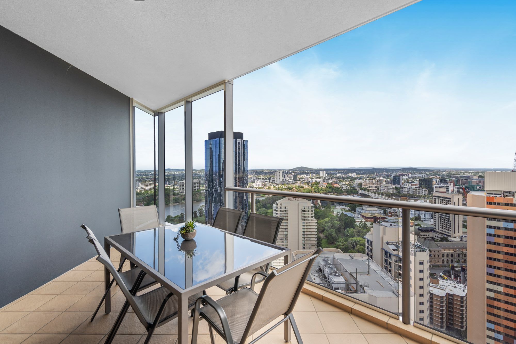 A balcony with a glass table and chairs, overlooking a cityscape with modern buildings.