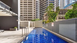 A swimming pool with a tiled floor, surrounded by a building with a geometric facade and a view of high-rise buildings in the background. - City Skyline from Level 37 | Free Parking, Pool & Gym (Brisbane City)