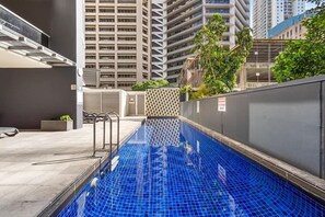 A swimming pool with a tiled floor, surrounded by a building with a geometric facade and a view of high-rise buildings in the background. - City Skyline from Level 37 | Free Parking, Pool & Gym (Brisbane City)