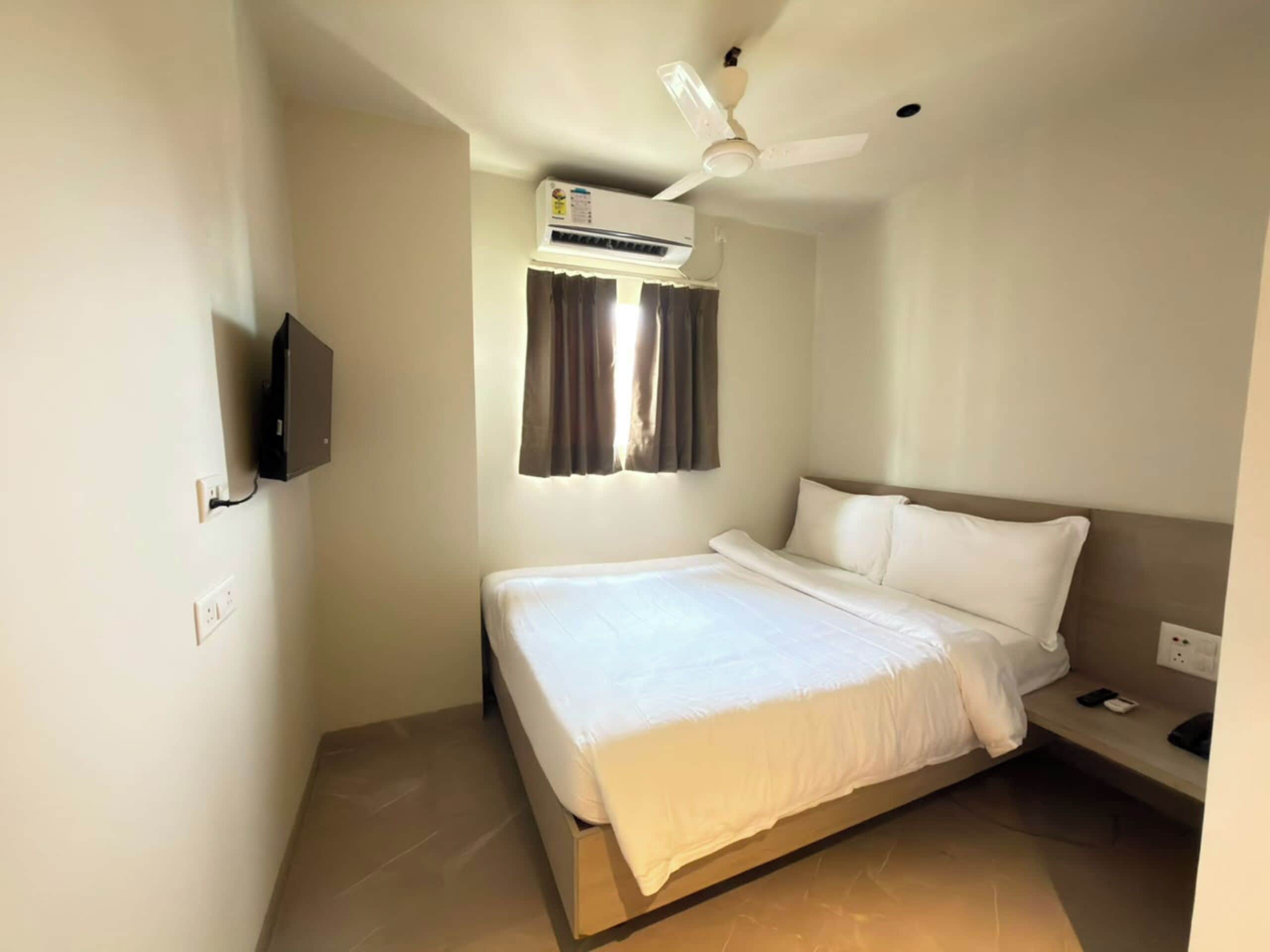 A hotel room with a bed, a ceiling fan, an air conditioning unit, a TV mounted on the wall, and a bedside table with a phone and a book.