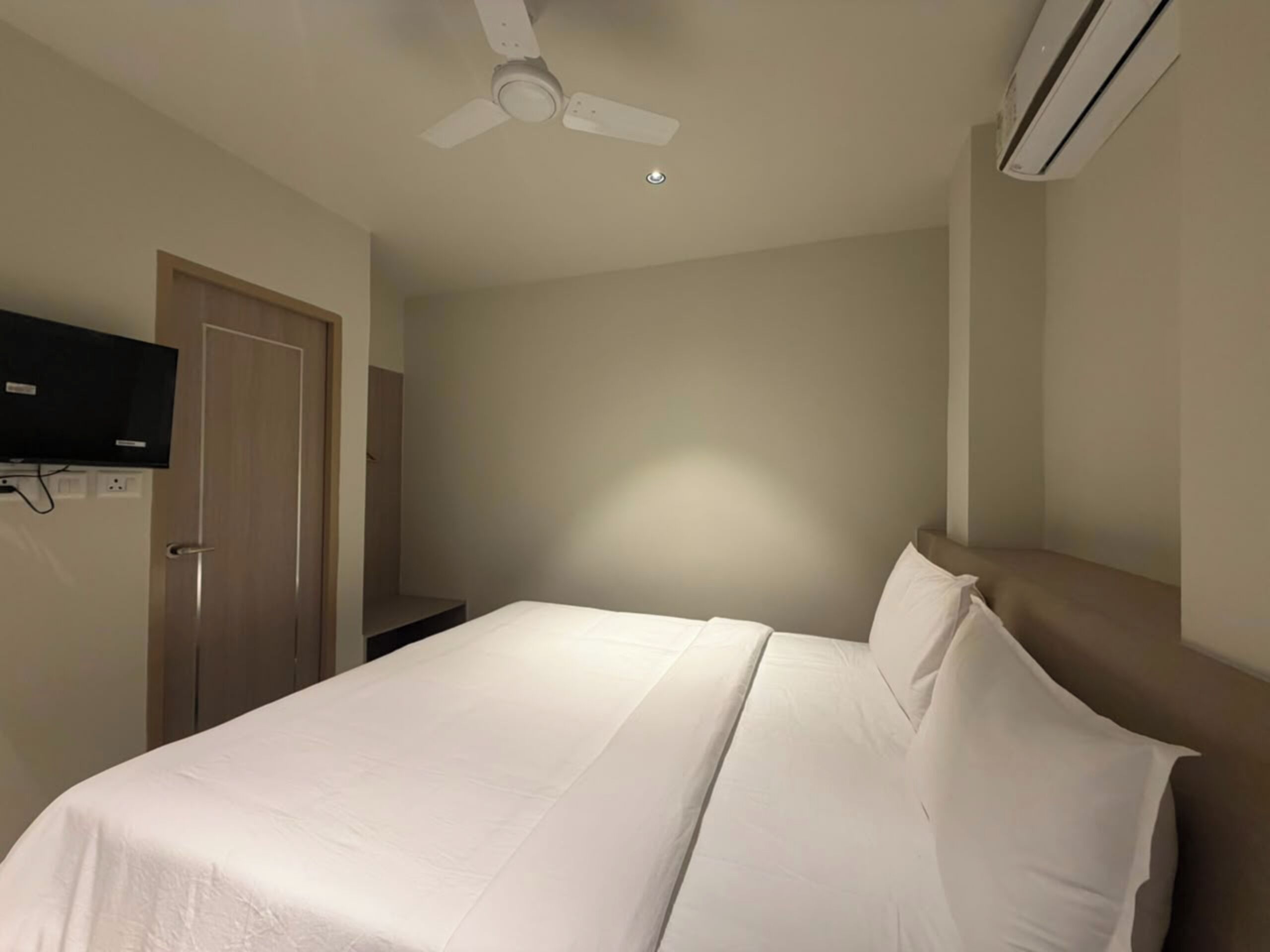 A hotel room with a bed, a ceiling fan, an air conditioning unit, and a television mounted on the wall.