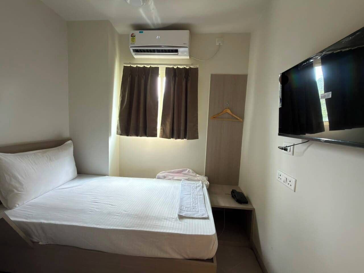 A single-bed room with a flat-screen TV, an air conditioner, a bedside table with a phone, and a window with curtains.