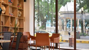 A hotel lobby with a large glass window, a wooden bookshelf with colorful items, and a seating area with chairs and a table.