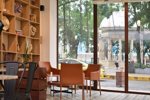 Un hall d’hôtel doté d’une grande baie vitrée, d’une bibliothèque en bois où sont exposés des objets colorés, et d’un espace détente avec des fauteuils et une table.