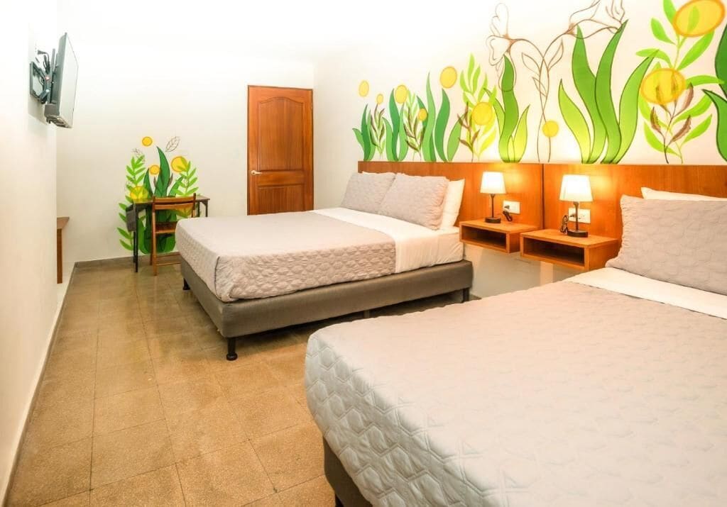 A hotel room with two beds, a wooden door, a small table with a plant, and a wall with a floral mural.
