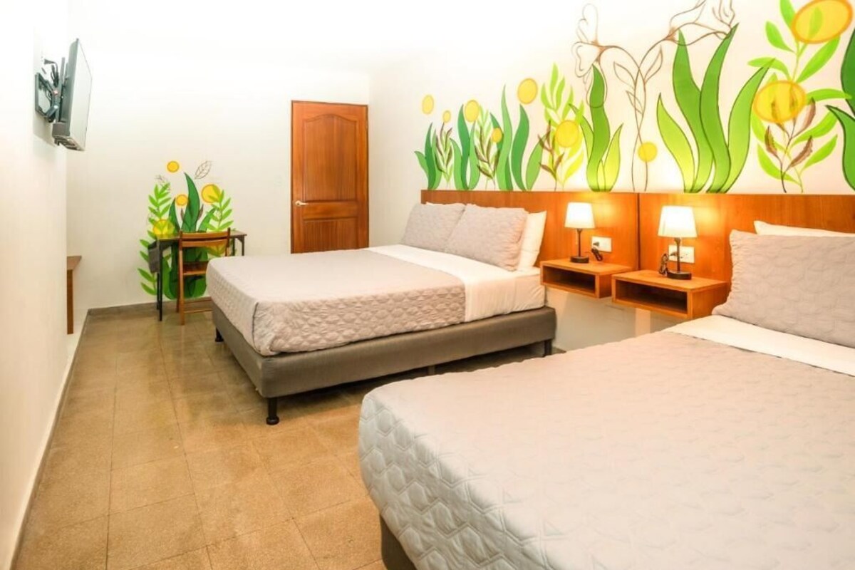Ein Hotelzimmer mit zwei Betten, einer Holztür, einem kleinen Tisch mit einer Pflanze und einer Wand mit floralem Wandbild.