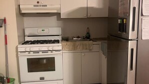 Een kleine keuken met witte kastjes, een gasfornuis, een roestvrijstalen koelkast en een groene emmer voor de mop.