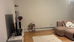 Een woonkamer met een houten vloer, een beige bank, een klein tafeltje met een vaas en een witte kast.
