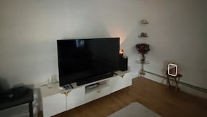 Een woonkamer met een flatscreen televisie bevestigd aan een witte kast, een klein tafeltje met een lamp en een stoel.