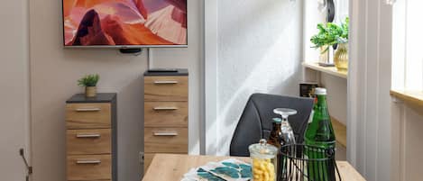 A modern dining area with a wooden table, a flat-screen TV mounted on the wall, and a window with curtains.