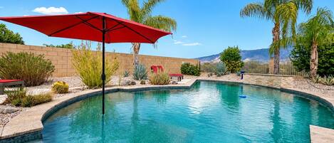 A swimming pool with a red umbrella, surrounded by palm trees and desert plants.