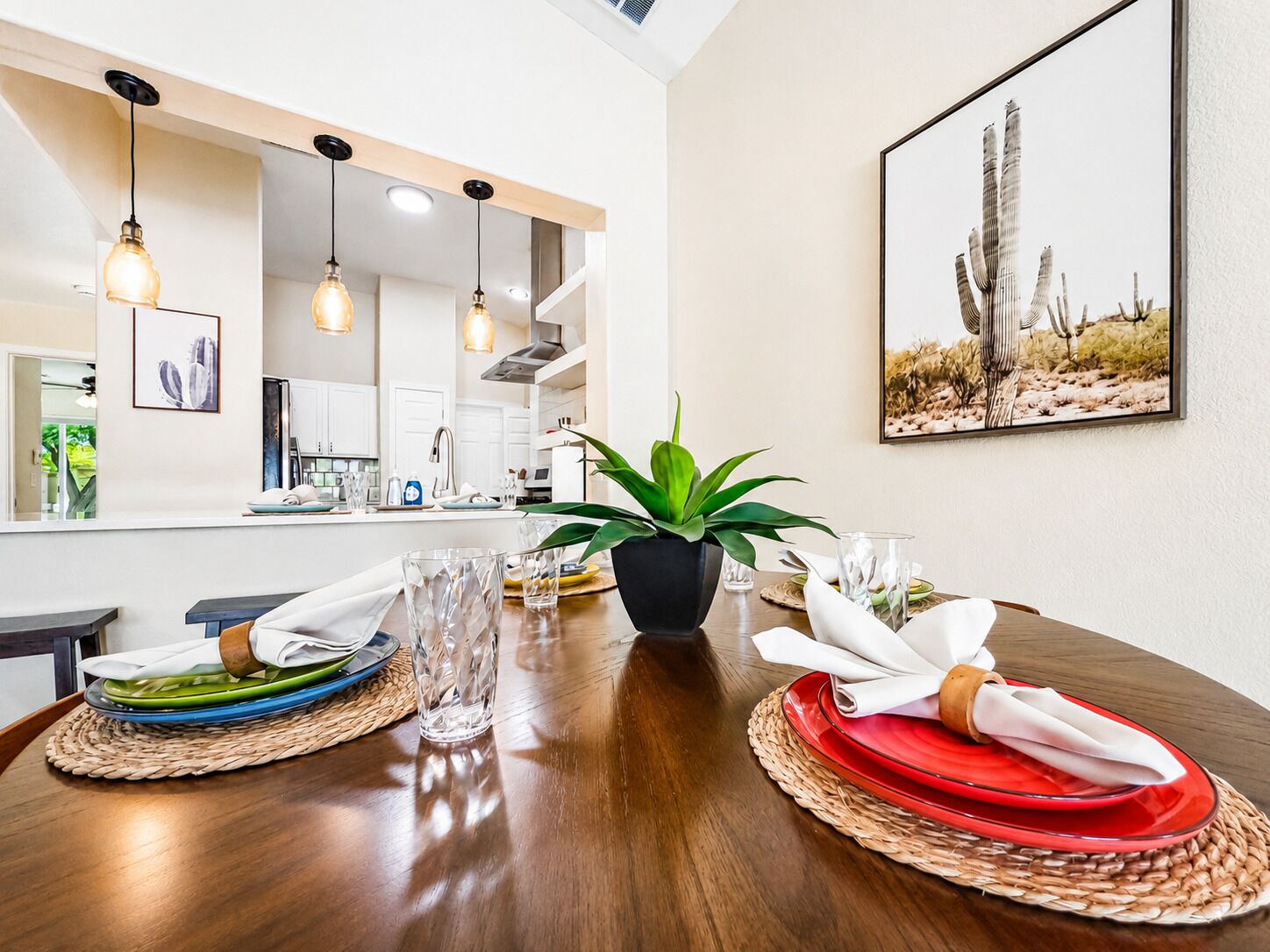 A dining area with a wooden table set for two, a potted plant, and a cactus-themed painting on the wall.