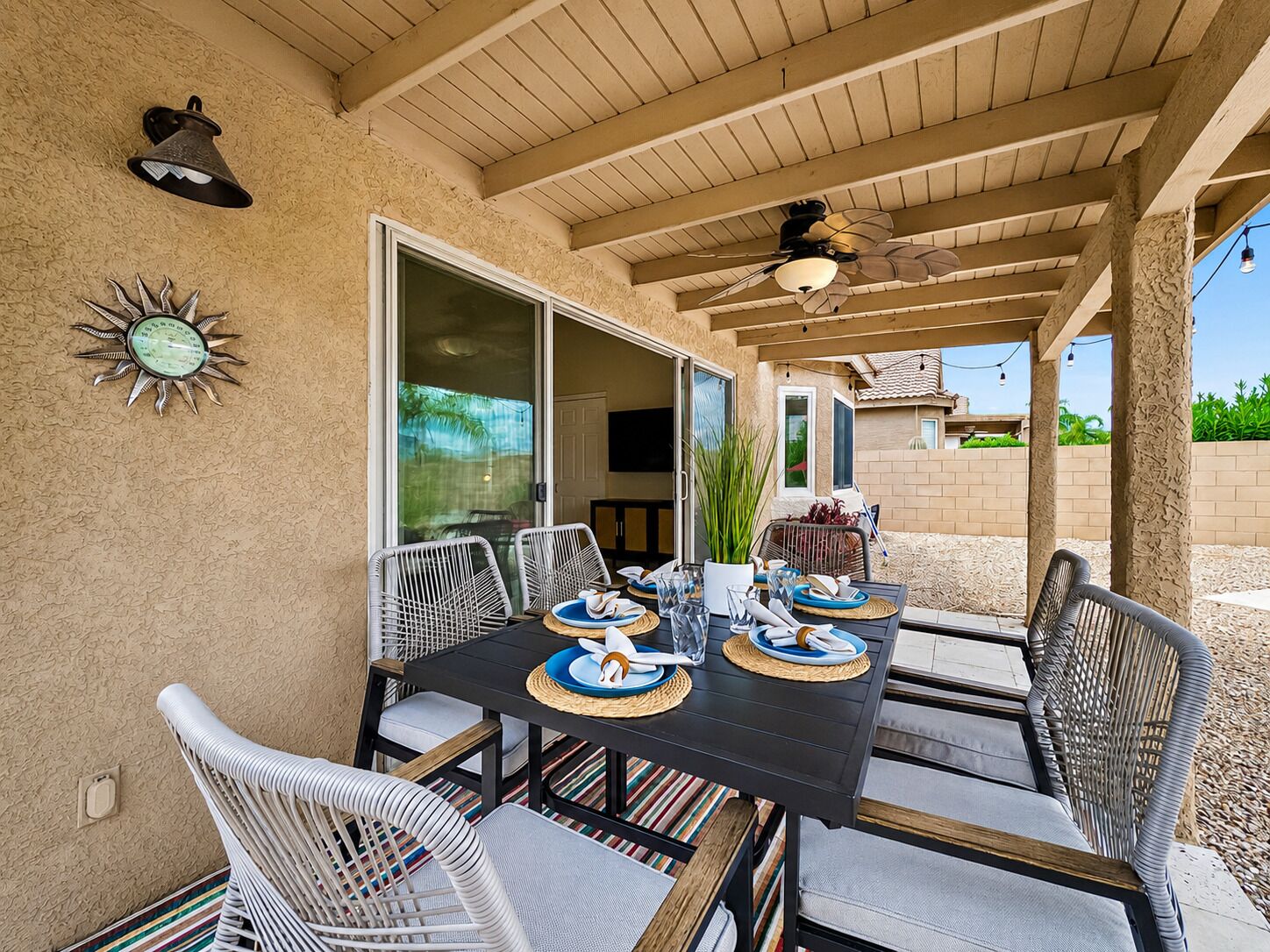 An outdoor patio with a table set for dining, surrounded by wicker chairs, a ceiling fan, and a decorative wall clock.