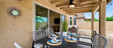 An outdoor patio with a table set for dining, surrounded by wicker chairs, a ceiling fan, and a decorative wall clock.