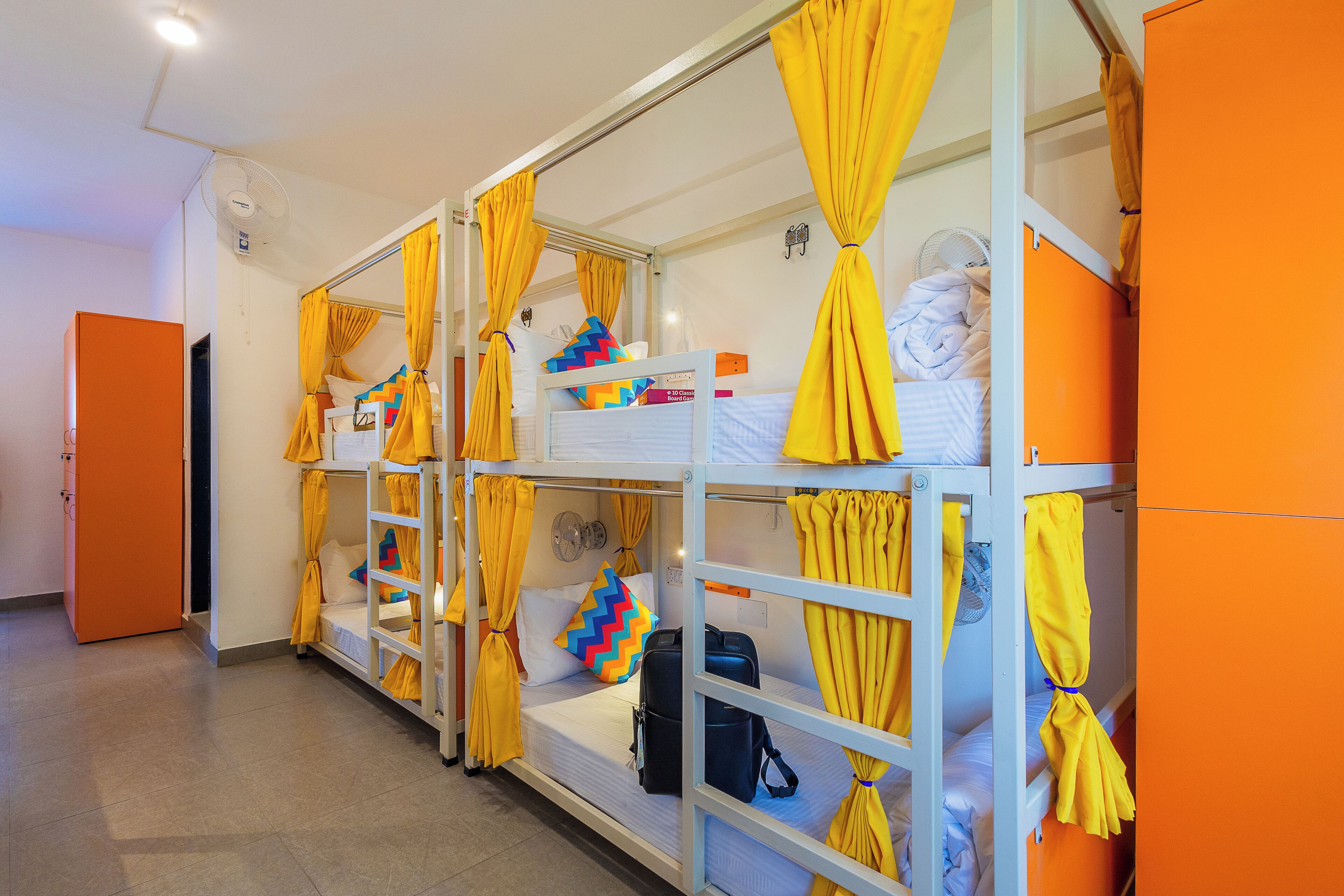 A dormitory room with bunk beds, each with yellow curtains, a white fan, and a small shelf with personal items.