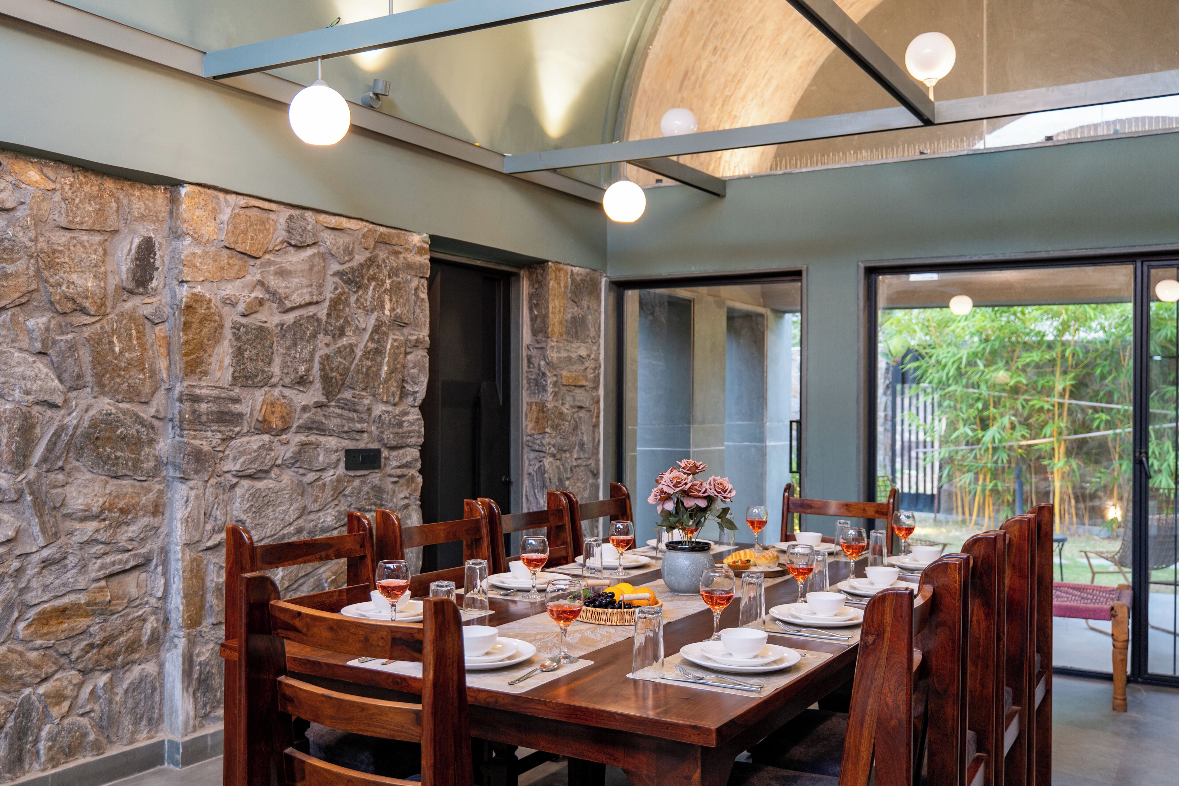 A dining area with a stone wall, wooden chairs, a long table set for a meal, and a view of greenery outside.
