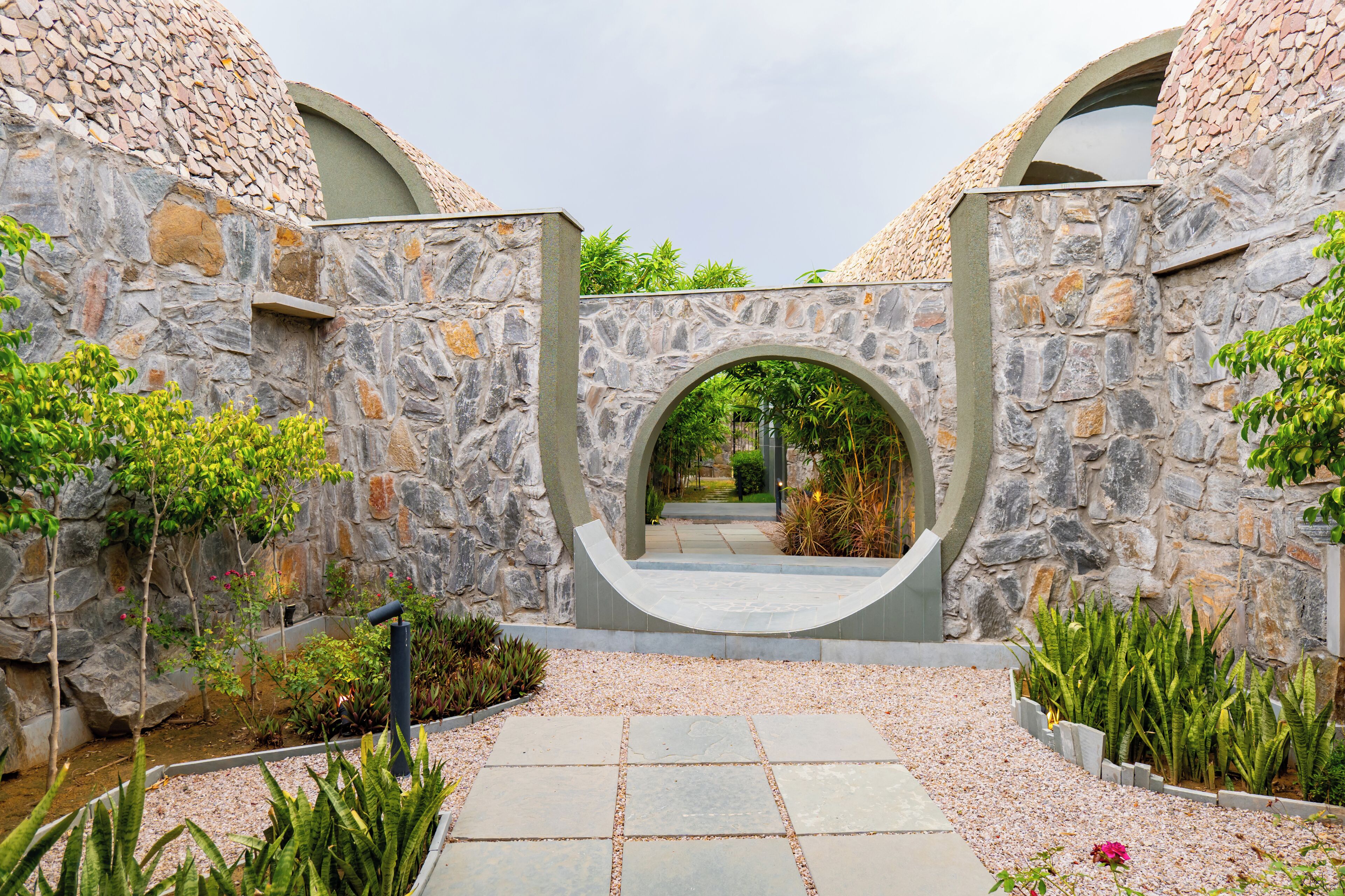 A stone archway leading to a courtyard with a pebble path and various plants.
