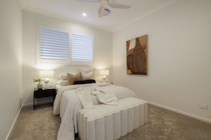 Un dormitorio con cama, mesitas de noche, ventilador de techo, ventana con persianas y un cuadro en la pared.