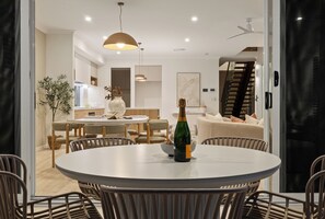 A modern dining area with a round table, chairs, a bottle of champagne, and a kitchen in the background. - Nordica – Stunning New Build, Bay Views, Steps to Wynnum & Manly Luxury Sleep 10 (Wynnum)