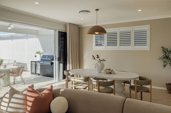 A modern living room with a sofa, dining table, and chairs, a grill outside, and a window with shutters.