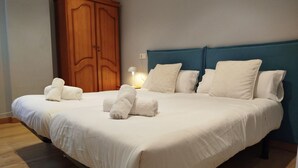 Een net opgemaakt bed met witte lakens en opgerolde handdoeken, een houten kledingkast en een nachtkastje met een lamp.