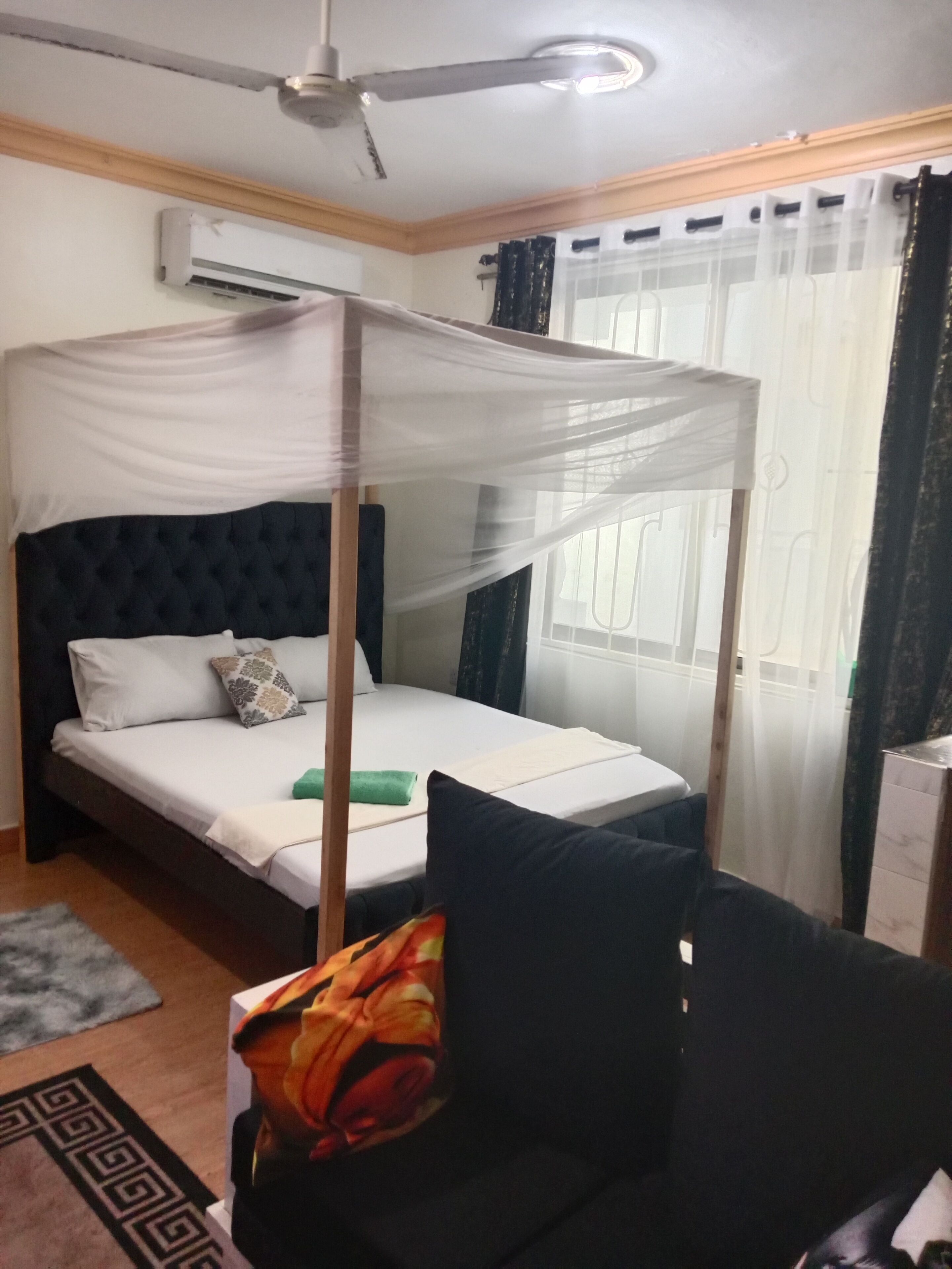 A bedroom with a four-poster bed, a ceiling fan, an air conditioning unit, and a window with curtains.