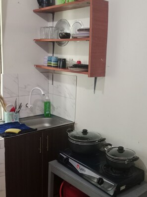 A kitchen with a sink, a stove, and shelves above holding dishes and a kettle. - Cozy studio apartment in quaint Mtwapa with an AC and Swimming Pool (Mtwapa)