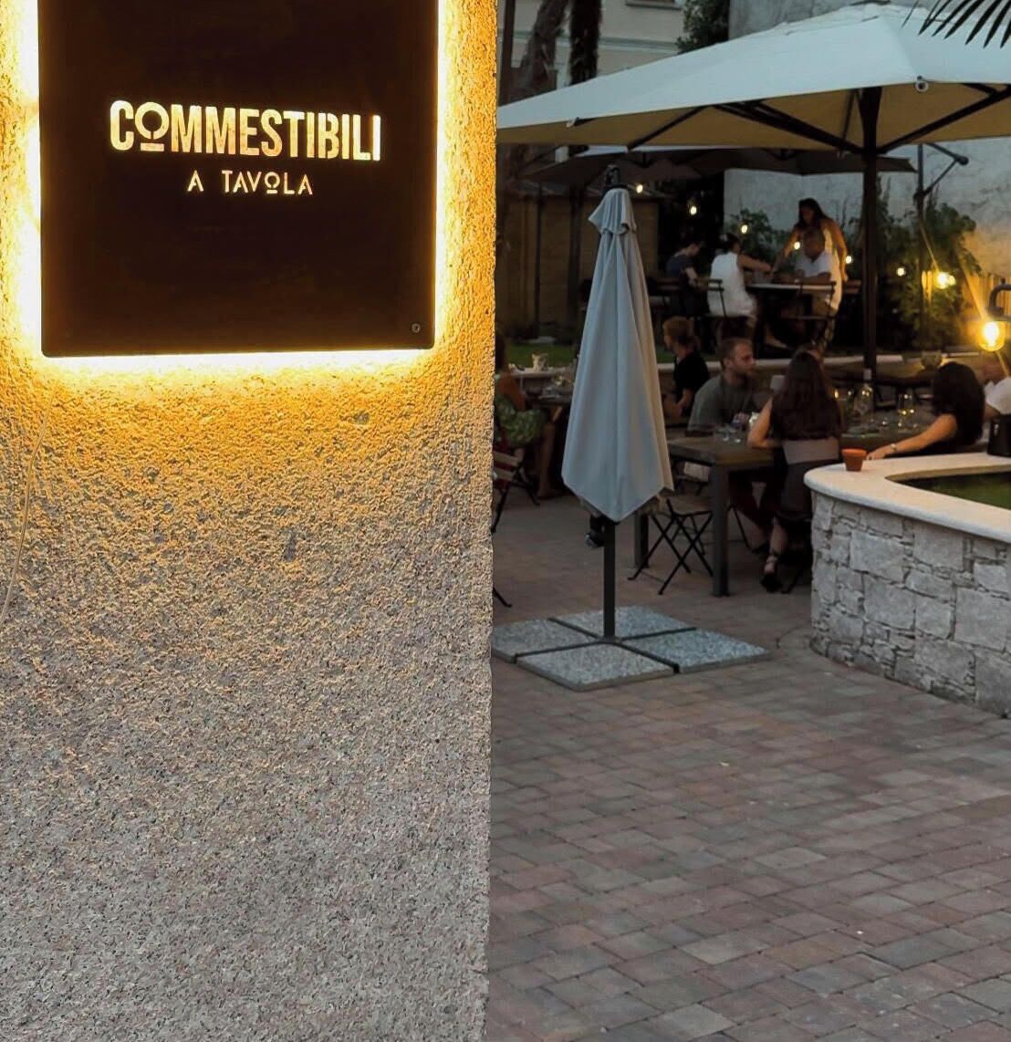 An outdoor dining area with tables and chairs, a sign with illuminated text, and a stone wall.