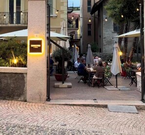 An outdoor dining area with tables and chairs, surrounded by buildings and potted plants. - Baveno Rose Apartment 3 (Baveno)