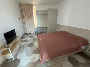 A hotel room with a bed, a TV on a wooden stand, a wardrobe, and a desk. - Baveno Rose Apartment 3 (Baveno)