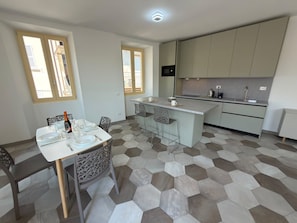 A modern kitchen with a central island, a dining area with a table and chairs, and a view through large windows. - Baveno Rose Apartment 3 (Baveno)