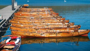 Una hilera de botes de remos de madera amarrados en un muelle, con un lago rodeado de bosque al fondo.