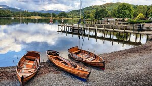Un lago tranquilo con barcas de madera en la orilla, un muelle que se adentra en el agua y montañas al fondo.
