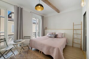 Un dormitorio con cama, una mesita con silla, ventana con cortinas y barandilla de balcón.