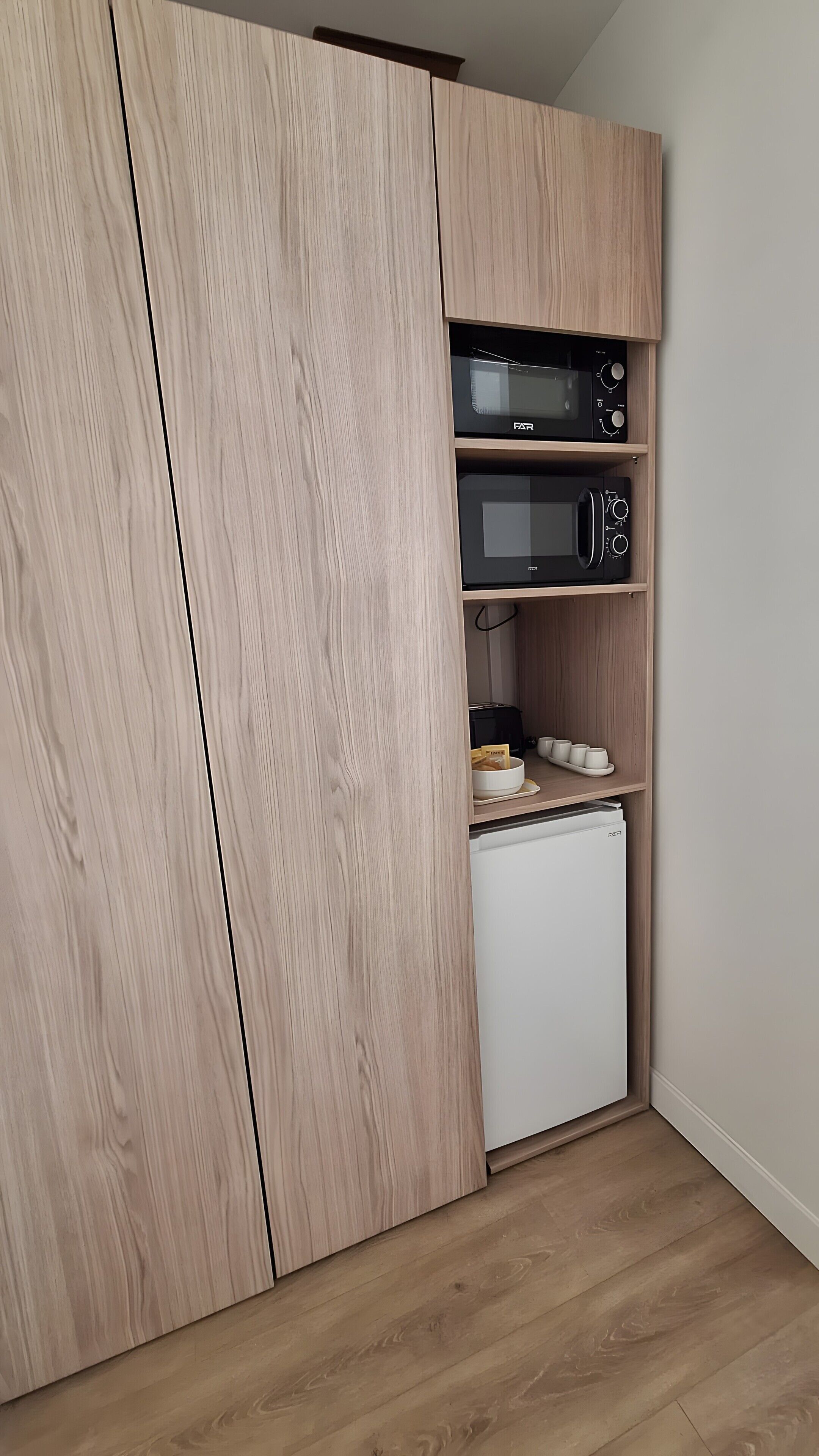 A kitchen with a built-in microwave, a small refrigerator, and a shelf with cups and a bowl.
