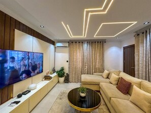 A modern living room with a flat-screen TV, a sofa, a coffee table, and a decorative ceiling light fixture. - 2-Bedroom Luxury Apartment | Fast Wi-Fi | Smart TV | 24/7 Power & Security (Lagos)