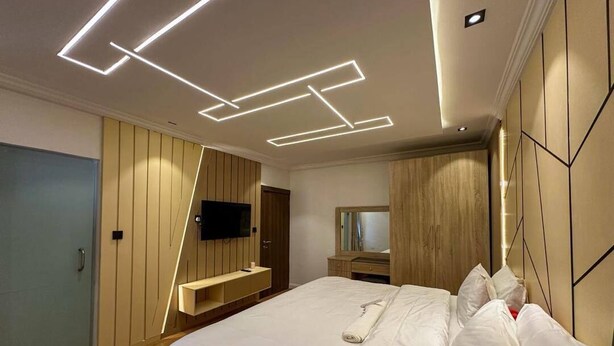 A modern bedroom with a bed, a flat-screen TV, a wooden wardrobe, and a mirror. - 2-Bedroom Luxury Apartment | Fast Wi-Fi | Smart TV | 24/7 Power & Security (Lagos)
