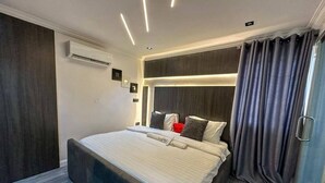 A modern hotel room with a large bed, dark curtains, and a built-in air conditioning unit. - 2-Bedroom Luxury Apartment | Fast Wi-Fi | Smart TV | 24/7 Power & Security (Lagos)