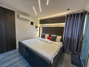 A modern hotel room with a large bed, dark curtains, and a built-in air conditioning unit. - 2-Bedroom Luxury Apartment | Fast Wi-Fi | Smart TV | 24/7 Power & Security (Lagos)