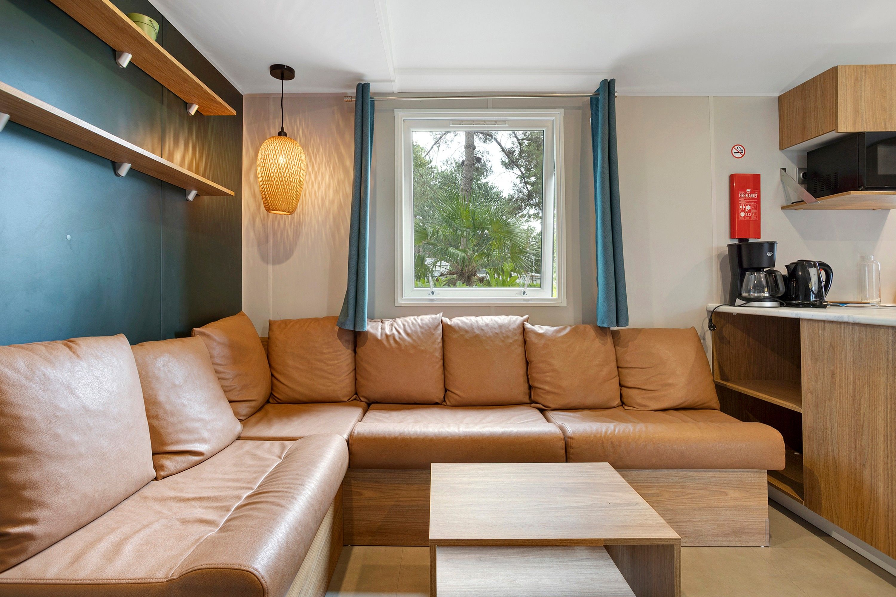 A modern living space with a leather sofa, a wooden coffee table, a hanging light fixture, a window with curtains, and a kitchenette area.