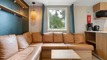 A modern living space with a leather sofa, a wooden coffee table, a hanging light fixture, a window with curtains, and a kitchenette area.