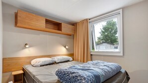 A bedroom with a bed, bedside tables, a built-in shelf, and a window with curtains. - Mobile Home “On Domaine Kerlann” with Shared Pools and Private Terrace (Pont-Aven)