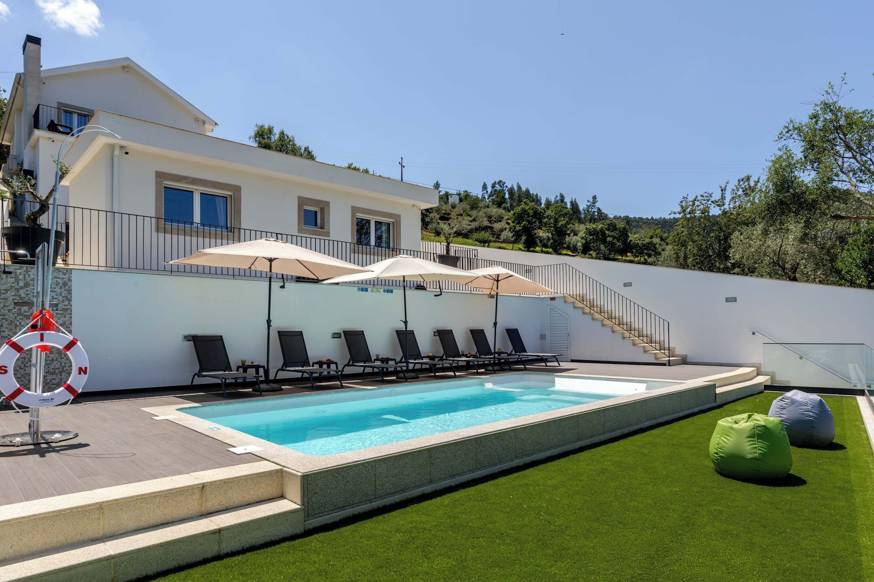 A modern pool area with a clear pool, sun loungers, umbrellas, and a staircase leading to a second floor.